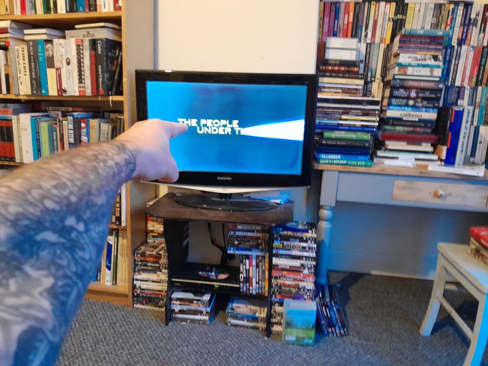 horrowfide's left arm pointing at the intro to the film people under the stairs on the television in his messy living room. 