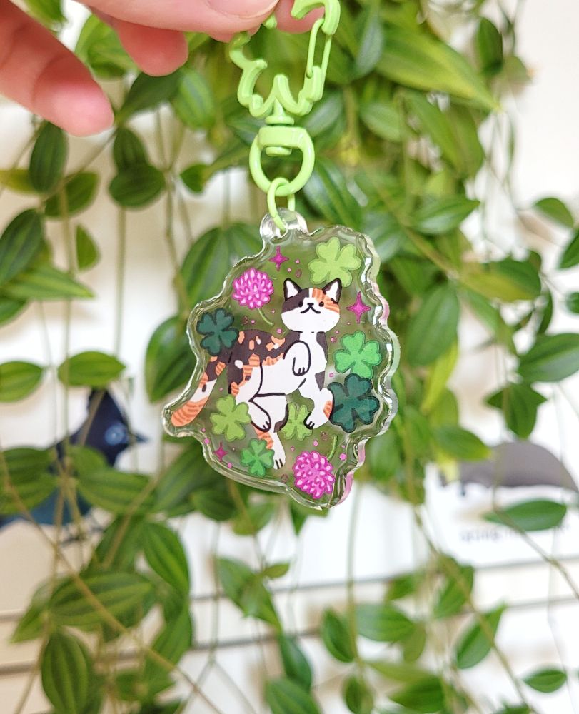 Photo of a keychain charm with a clover shaped keyclasp. The design is of a calico cat surrounded by clover leafs and flowers. The background of the photo is a house plant.