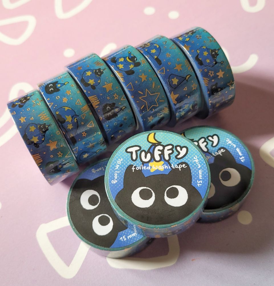 Photo a a stack of washi tapes. The motive is my wizard cat OC Tuffy a small black kitten with a blue pointy wizard hat and a cloak. The washi tapes has mainly blue colours and stars in gold foil.