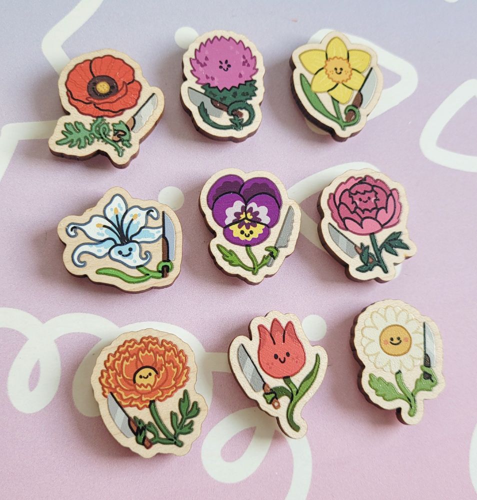 Photo of 9 flower magnets in cartoony style. They all have little faces and are holding a knife.
Row 1 from left to right: poppy, thistle, daffodil
Row 2 from left to right: lily, pansy, peony
Row 3 from left to right: marigold, tulip, daisy