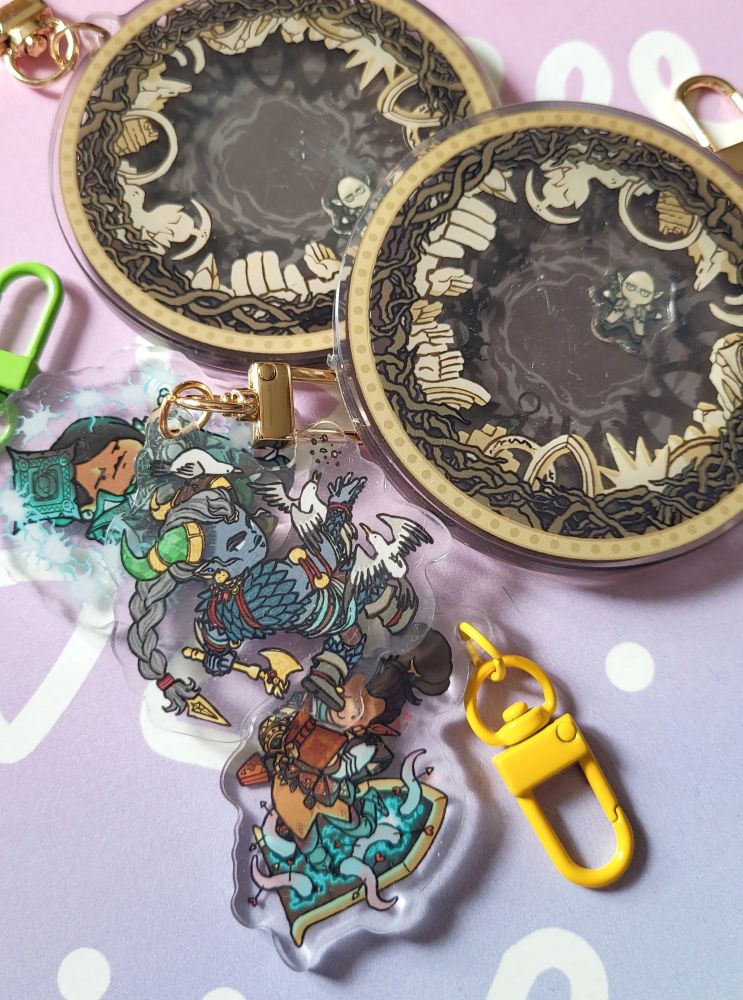 Photo of a small pile of acrylic charms of Dragon Age The Veilguard characters Bellara, Taash and Neve. The big round charm at the top is a shaker charm of (tiny) Solas trapped in regret prison.
