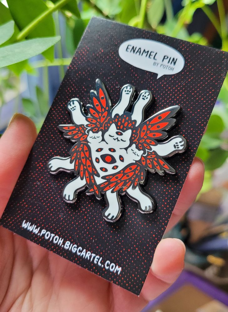 Photo of my hand holding an enamel pin on a backing card. The pin is a biblically accurate cat in white, with 3 heads and 6 wings. Inside the wings and on the cat are countless red eyes. Stretched out in a star-like fashion are the 8 legs of the cat.