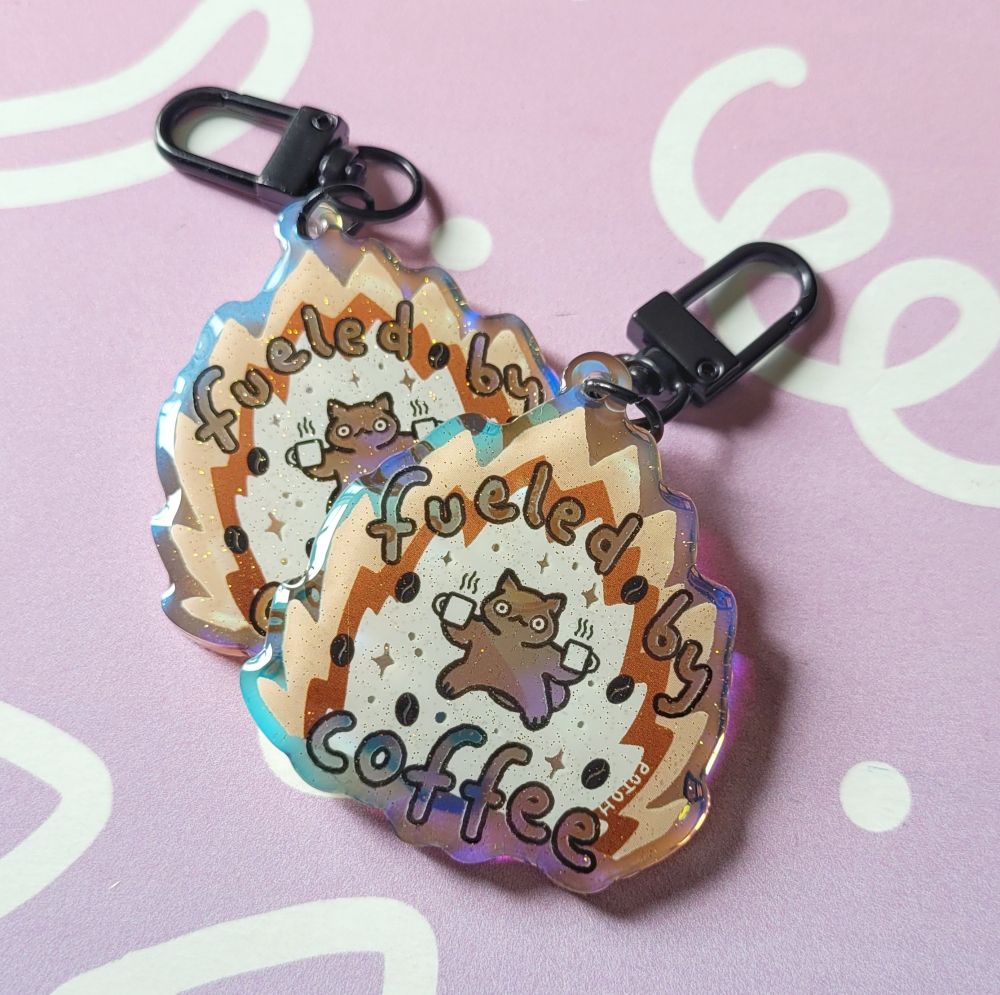 Photo of 2 holo glitter charms. The motive is a small cat in a wide stance and wide stare holding a coffee cup in each hand. The cat is surrounded by brown flames with the words "fueled by coffee" in them.