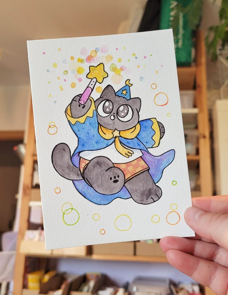 Photo of a drawing of my wizard cat OC Tuffy. He's wearing his pointy wizard hat, cloak and shorts. He's jumping up, looking up to is star wand he is holding up. Colourful circles are sprinkled around the wand and the rest of the page. The drawing is coloured with watercolour paint.