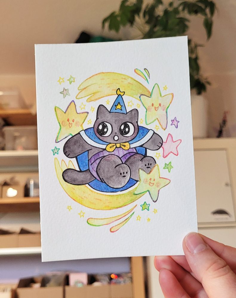 Photo of a drawing of my wizard cat OC Tuffy. He's wearing his pointy wizard hat, cloak and shorts and is circled by shooting stars. The drawing is coloured with watercolour paint. The shooting stars are outlined with fineliners in different colours for a rainbow effect.