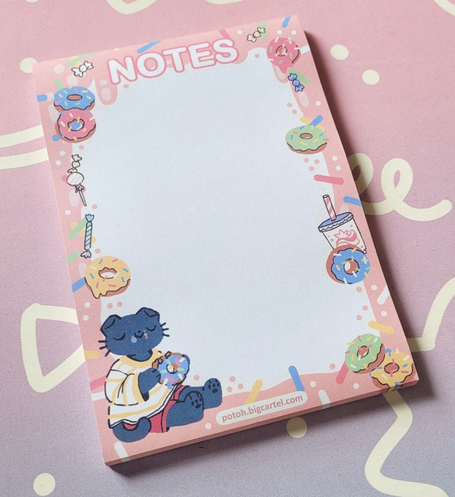 Photo of a blank paper notepad with a pink donut pattern as a border and a cat sitting in the bottom left corner eating a donut.