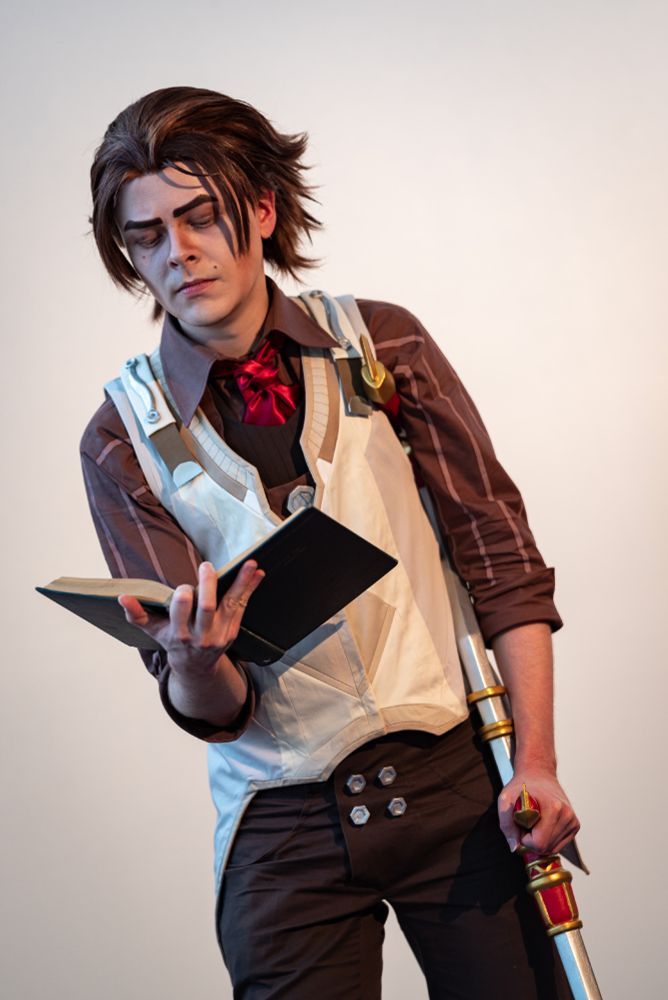 Picture of me in Viktor cosplay from Arcane. The costume is the version from season 1 act 2. I am standing and reading a book, with a stern expression on my face. The background is just a white wall. The photo is cropped above knees.
