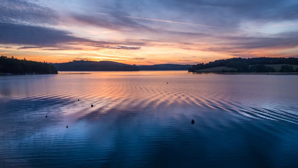 A peaceful sunrise over Lake Vassivière, Auphelle, France. Gentle ripples on the lake are lit in gold by the rising sun between and in front of low dark hills. Blue-grey clouds are reflected in the water. 