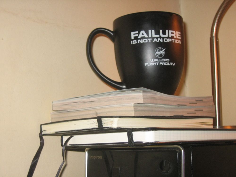 A photograph of a black mug with a long thin shaped handle that boldly states "FAILURE" in large white text on top of the second line of text that is slightly smaller and states, "IS NOT AN OPTION". Centered below that statement is the NASA logo in pure white, and below that white text states repeating the two line format breakdown but in a smaller and softer edged font that some would say seems rather space tech friendly, "WALLOPS" on top of "FLIGHT FACILITY". The mug rests upon a stack of two softcover books and two hardbound notebooks with black ribbon for page markers and bands to hold them together. None of the spines are facing the camera and so their contents are a mystery. Next to both is a brushed silvery metal base of a desk lamp with a gooseneck portion that reaches off frame so the shade cannot be seen. Below all of that is the top of a computer tower that is black with some silver edging. In the upper left corner of the tower the word "Inspiron" can be seen.