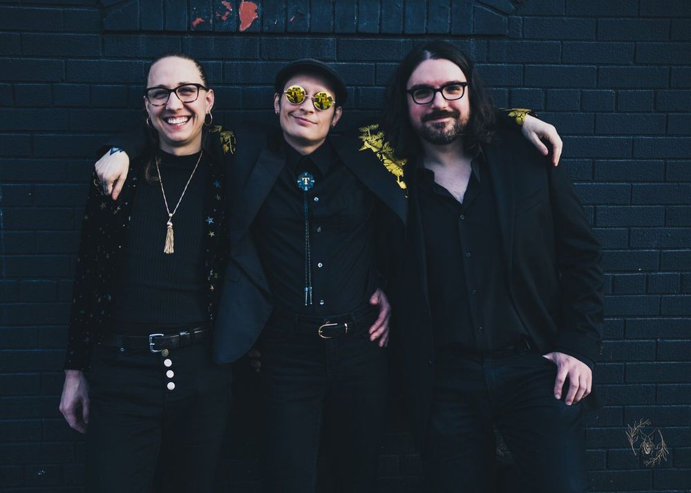 Three smiling people wearing black stand with their arms around each other 