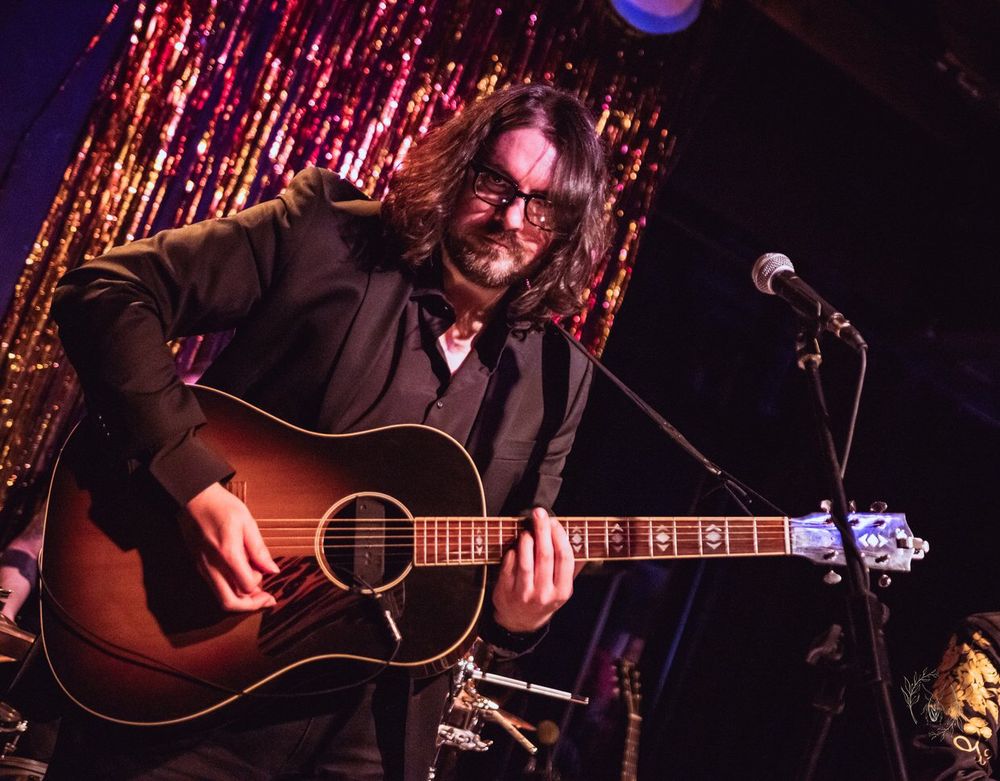 A man wearing all black glares at the camera as he plays a guitar. He has glasses and dark, shoulder-length hair.