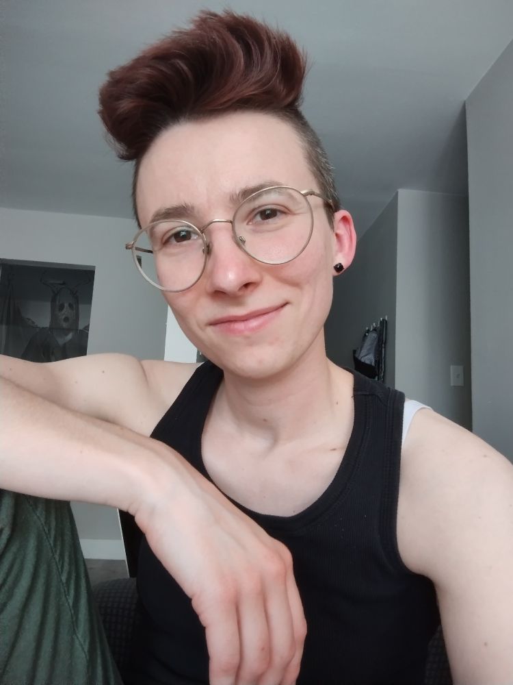 A butch woman poses for a selfie. She's white and has a wide mohawk dyed an auburn color. She's wearing a blank tank top and stud earrings. In the background, if you look really closely, there's the faint outline of something demonic lurking in the open doorway behind her... wait... wait, what IS that? It has a pale face, black eyes and a wide gaping mouth. Are those... wings? It's so faint, you almost can't make it out. Is it a ghost? Is she haunted?? Will anyone notice this and point it out?