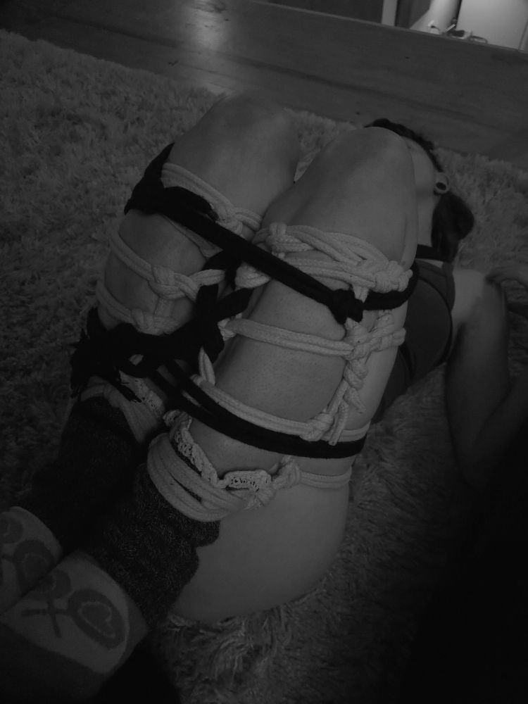 a woman lying on her back on a shag rug. her legs are tied in black and gray (pink) rope, in futomomo ties. her legs are covering her chest and face. the photo is monochrome and taken from a slight side angle. 
