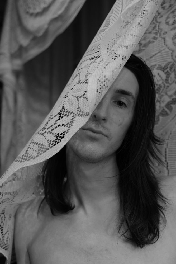 puppii from the chest up with lace in front of the right side of their face. their hair is falling onto their shoulders. the photo is monochrome 