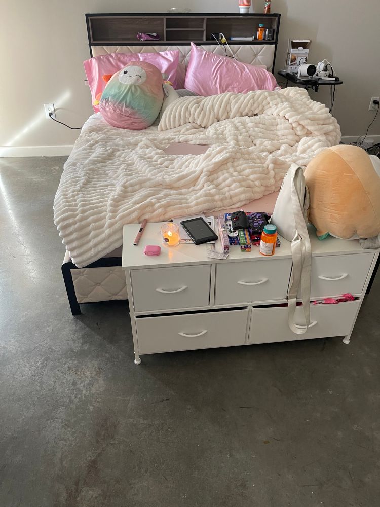 Picture of a queen sized bed with a bed frame that includes a headboard with storage space and outlets/usb ports. On the bed is a fluffy cream colored comforter/blanket, two sets of pillows, and two Squishmallows. In front of the bed is a small dresser with a variety of items on it and a large Rowlet plush.