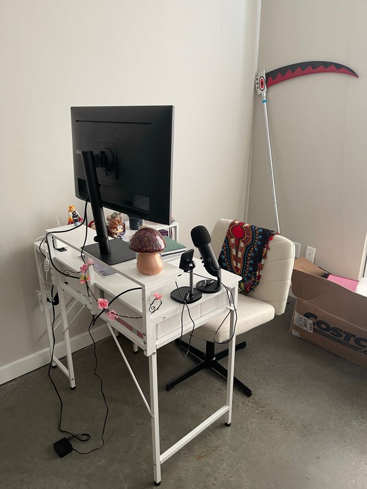 Pictured is an L-shaped desk with a large screen on the upper shelf, a large office chair (one of those viral criss-cross chairs), various items decorating the desk top, and a cosplay prop: Maka’s scythe from Soul Eater 