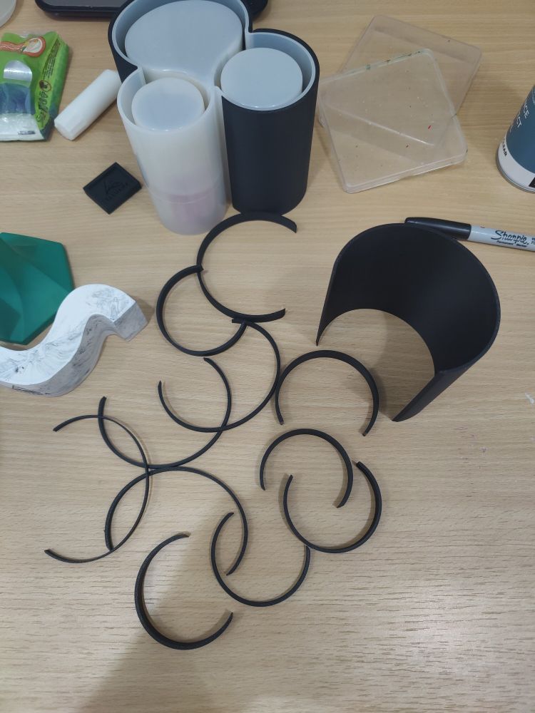 A bunch of roughly semi-circular plastic strips strewn on a desk alongside a much taller curved piece.
Near them is a silicone mould with two similar plastic curves acting as supportive shells around parts of the mould. A third of the mould has no support.
Some other crafting junk is also on the table.