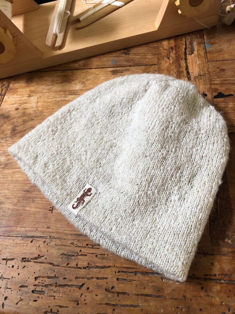 A textured white knit hat with a little woven tag, featuring a newt. There is a rigid heddle loom in the background. 