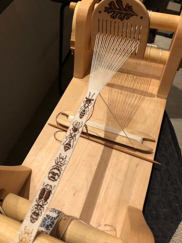 A woven band featuring small ornate images of four different ant species. The background is white and the ants are woven in brown. The band is being woven on a homemade loom with an oak leaf motif. 