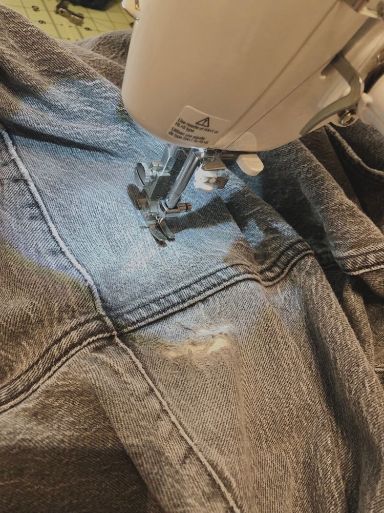 A gray pair of jeans with holes in the inner thigh, being repaired with machine darning 