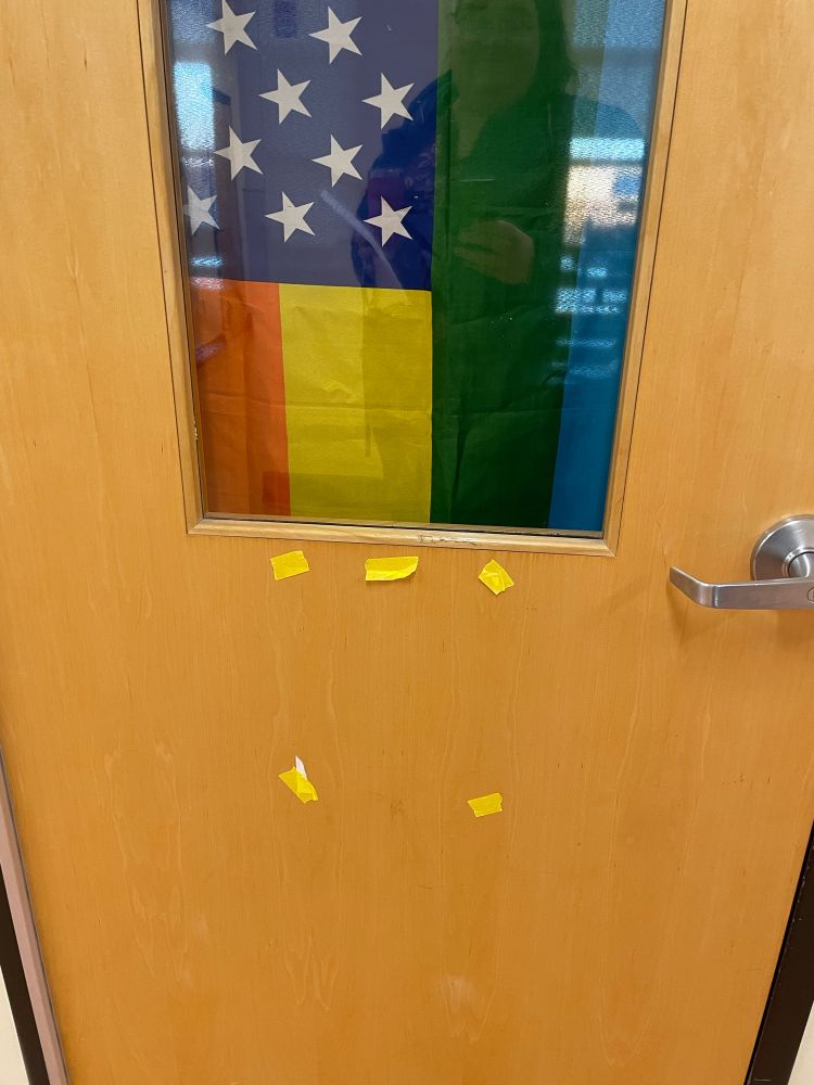 Classroom door with club poster torn off. Rainbow American flag is visible in the classroom. 