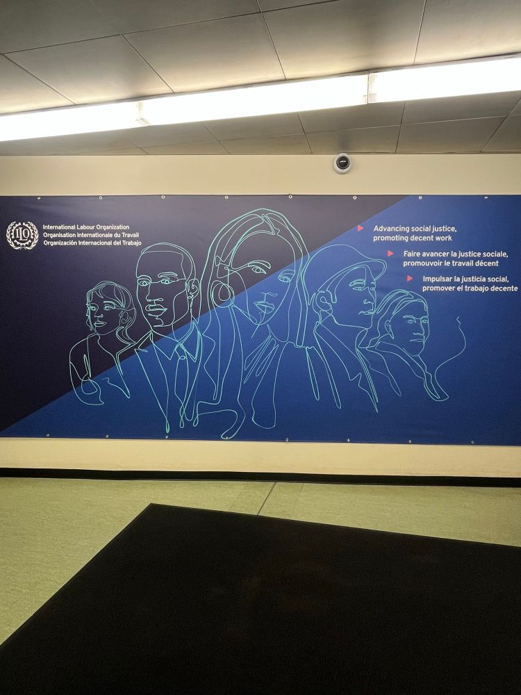 A board on a wall with the ILO emblem, a drawing of 5 people and a phrase in English, French and Spanish.