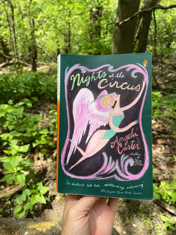 A person holds a paperback copy of Nights at the Circus by Angela Carter, trees in background, while in a forest during a quick break from windowless basement work