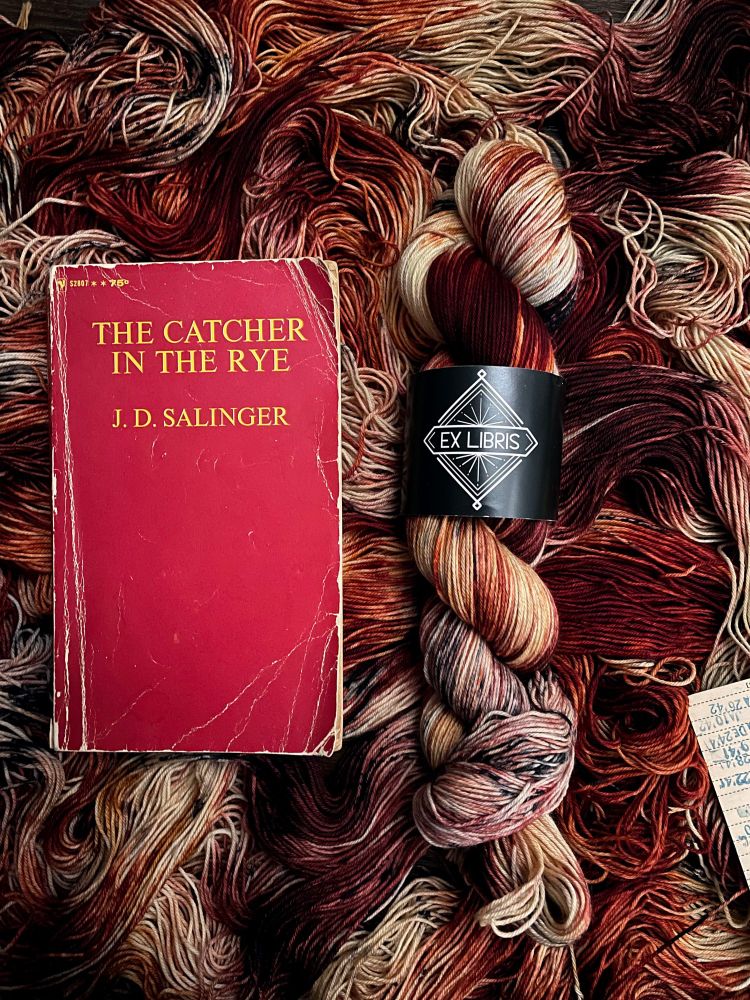 A skein of dramatic dark red and paper colored yarn with black speckles sits next to a copy of The Catcher in the Rye. Part of Ex Libris Fibers Indelible monthly limited color fundraising yarn series; a percentage of sales benefit ALA's Unite Against Book Bans & the Queer Liberation Library