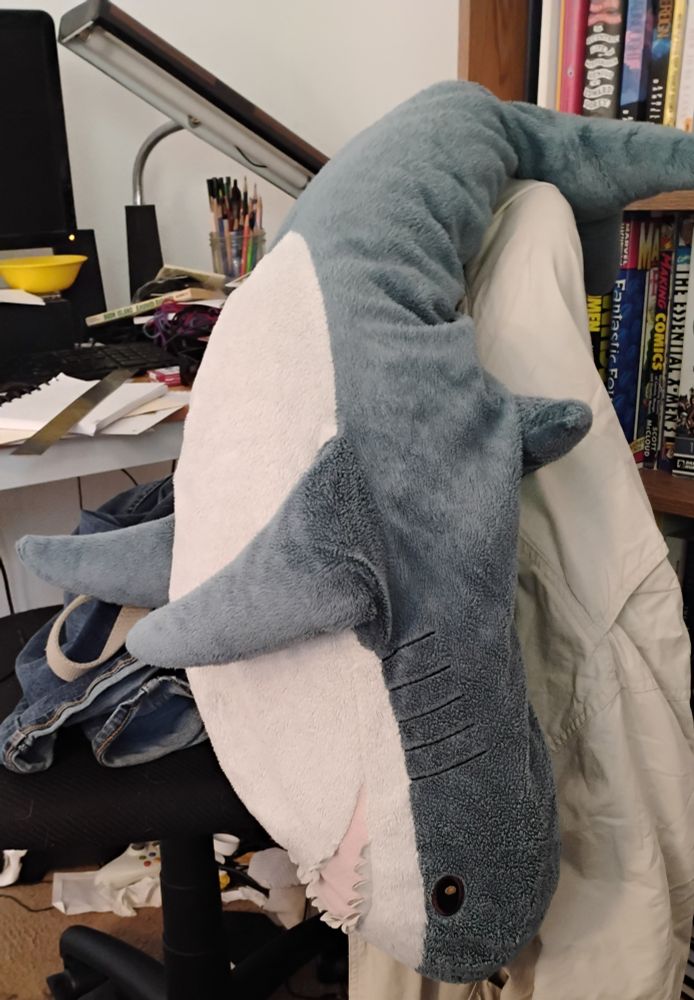 A blåhaj hanging upside down on the back of an office chair