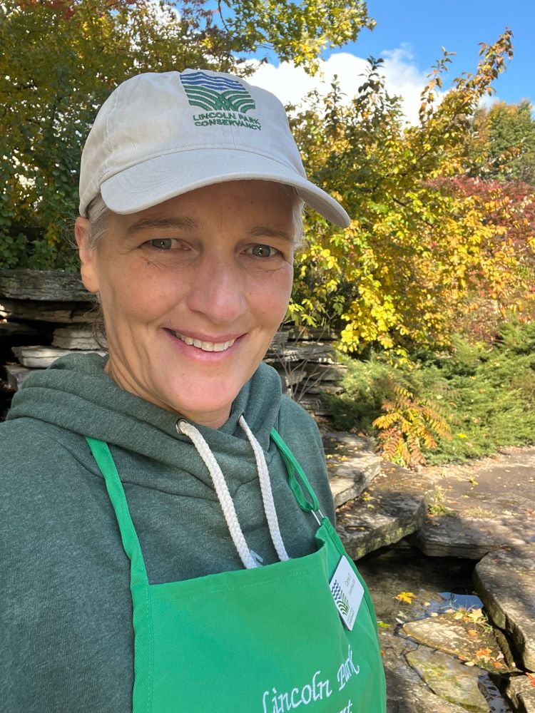 Selfie of me with fall foliage in the background. I’m wearing a baseball cap, green hoodie, and bright green apron that says Lincoln Park Docent
