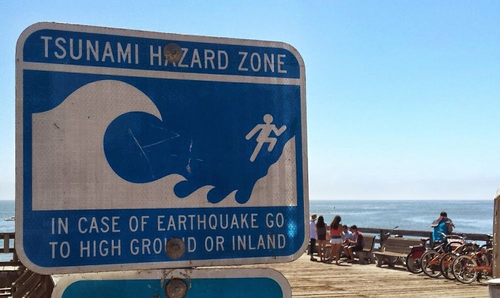 A blue and white "Tsunami Hazard Zone" sign posted on the wooden deck of a wharf. The sign includes an image of a stylized large wave and a stick figure fleeing up a hillside, and the text "In case of earthquake go to high ground or inland." In the background, we see people leaning against wooden railing and, beyond that, the blue waters of the Pacific Ocean on a sunny day. Seveal bicycles are also seen on the railing.