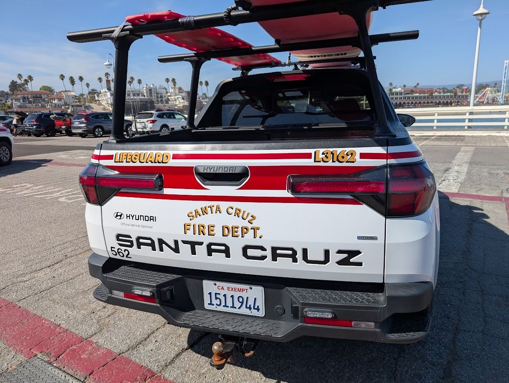 Back of a Hyundai Santa Cruz used as a lifeguard truck with the logo of the Santa Cruz Fire Department. A truck rack over the bed holds two surfboards. The photo was taken from the Santa Cruz Wharf lifeguard station.