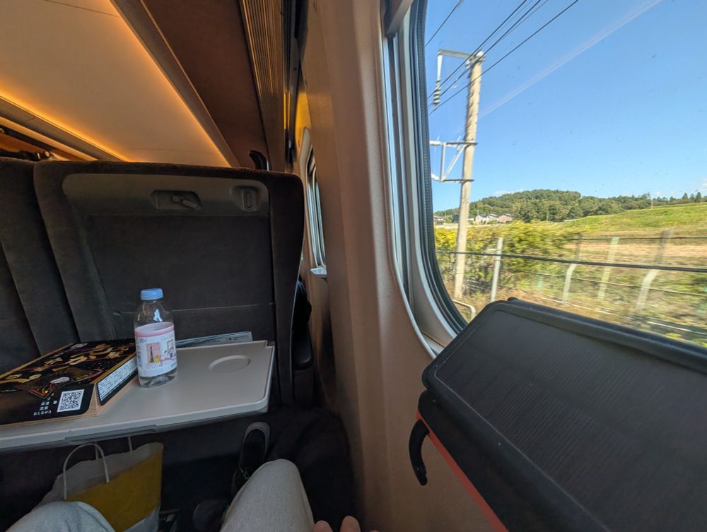 The view from my seat on the Hayabusa Shinkansen E5 train. On the tray table in front of my seat I have an ebiben box lunch and a bottled water. Farmland is visible through the window on my right.