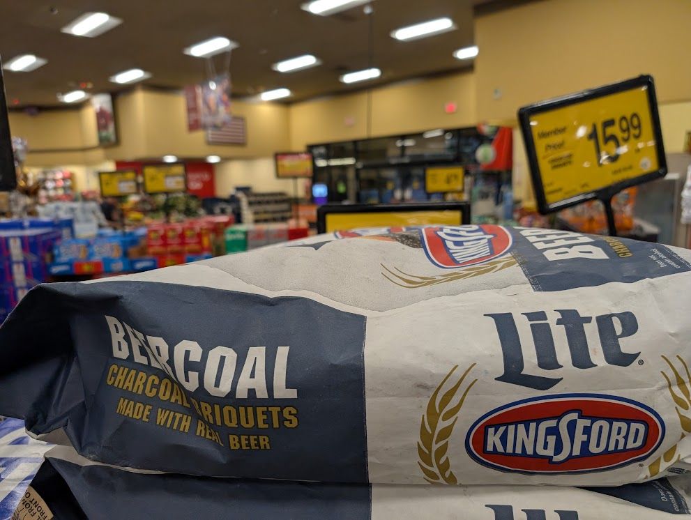 The side of a bag of Kingsford barbecue charcoal briquets with the Miller Lite logo above the Kingsford logo, and "BEERCOAL charcoal briquets Made with Real Beer" printed on the bag. Beyond the bag we can see grocery store signs and displays.