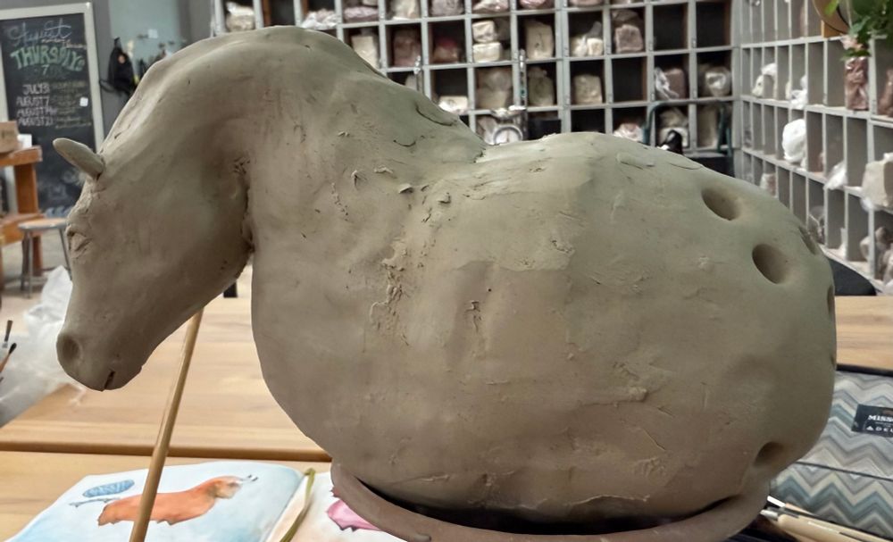 Stylized beige horse in clay sitting on a terra cotta stand surrounded by ceramics paraphernalia. The background is a busy studio.