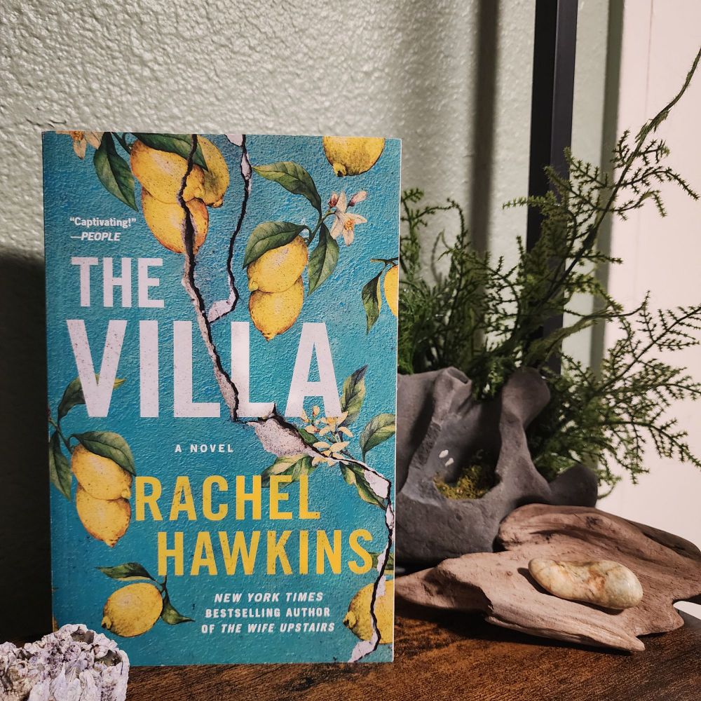 The villa by Rachel Hawkins sits on a shelf. The Book cover has a lemon tree in fruit against a teal concrete building