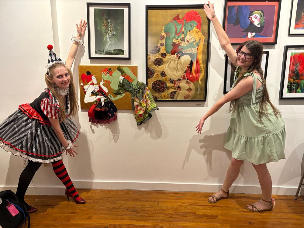 A photograph of two people in showleft showright poses, to the left and right of a multimedia painting of Ivy cheekily leaning in to kiss Harley, who has her fingers on Ivy’s lips (their dresses are fabric patches sewn together) and an illustration, heavily inspired by Klimt’s “The Kiss,” of Poison Ivy, draped in a floral-patterned dress, kissing the temple of Harley Quinn, who is wearing a ruffled collar and a leotard that is patterned in card suits.