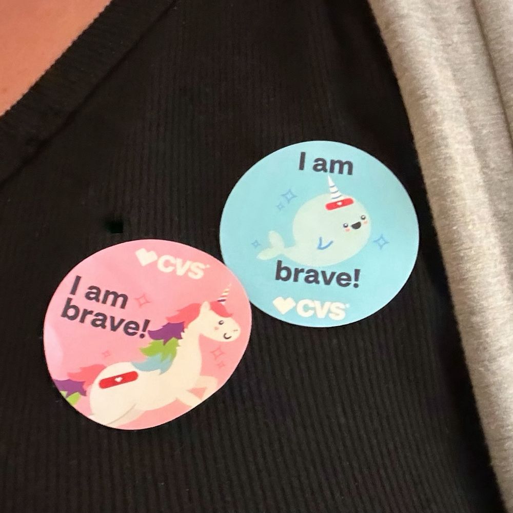 A close up of two stickers on a shirt. Both say “I am brave!” One is pink with a cartoon unicorn with a red bandage and the other is blue with a cartoon narwhal with a red bandage.