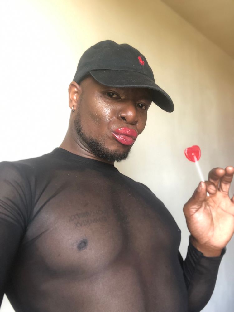 portrait photo where I am made up I am wearing a Ralph Lauren cap and a sheer you can see my nipple piercing, I have a duck mouth and I am holding a heart-shaped lollipop in my left 