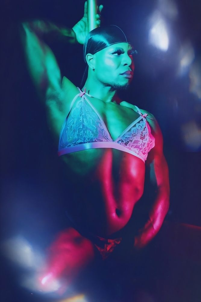 I'm squatting in front of a pole dance bar, there are green lights, I'm wearing makeup, a black durag, a pink lingerie set and very high black heels, I'm dancing lasciviously in front of the photographer.