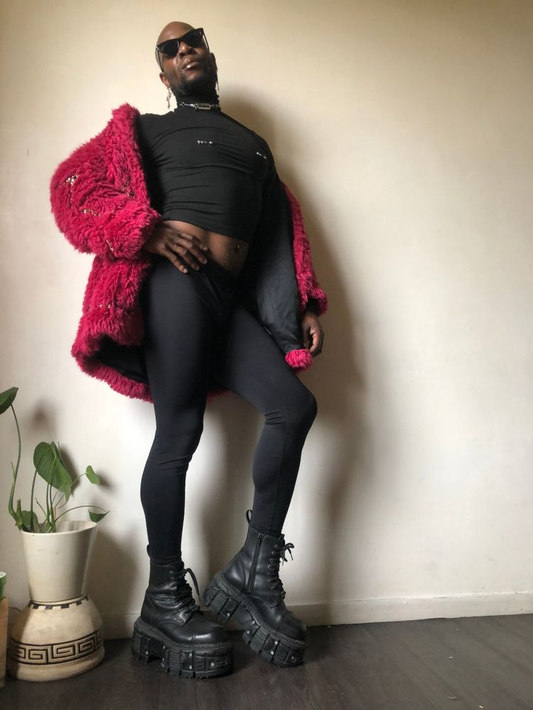 I am in profile, I am holding my hip with my right hand, my head slightly tilted back, I am wearing black leggings and high platform leather boots from New Rock, a long-sleeved black crop top with charms like nipple piercings, a vegan fur coat decorated with a fuchsia pink gold chain from Moschino and vintage sunglasses.
