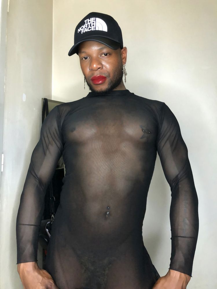 I am standing, I am wearing a transparent black t-shirt, you can see my navel piercing, and my nipple piercing with a star around it, I am wearing lipstick and gothic earrings, an adjustable cap on my head, I am pulling my t-shirt down with both hands.