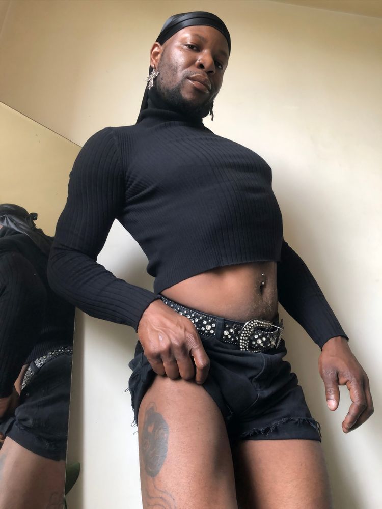 I am standing slightly arched, staring into the camera lens while gripping my shorts, I am wearing a black long-sleeved crop top sweater, my tattoo of my own face wearing a latex mask with a ponytail, my navel piercing is visible and I am wearing a durag and gothic cross earrings