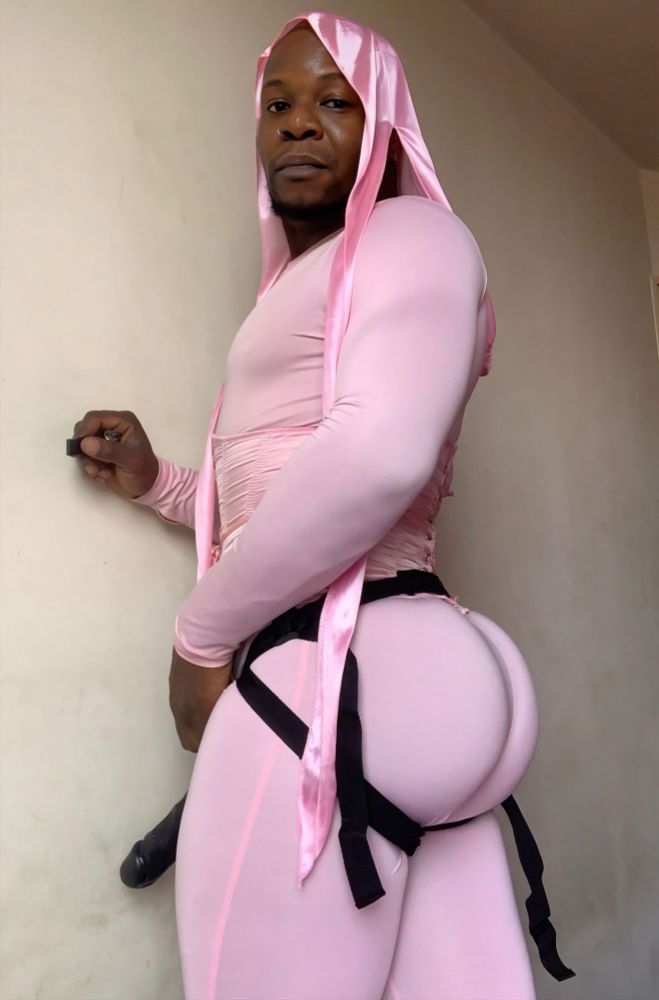 I am standing in profile, staring at the camera, I am wearing a very tight pink jumpsuit with a corset of the same color and a pink durag too, I am wearing a black strap-on dildo with a big black veiny cock.
I have my right hand on the wall in front of me, I love my ass if only I could fuck myself.
