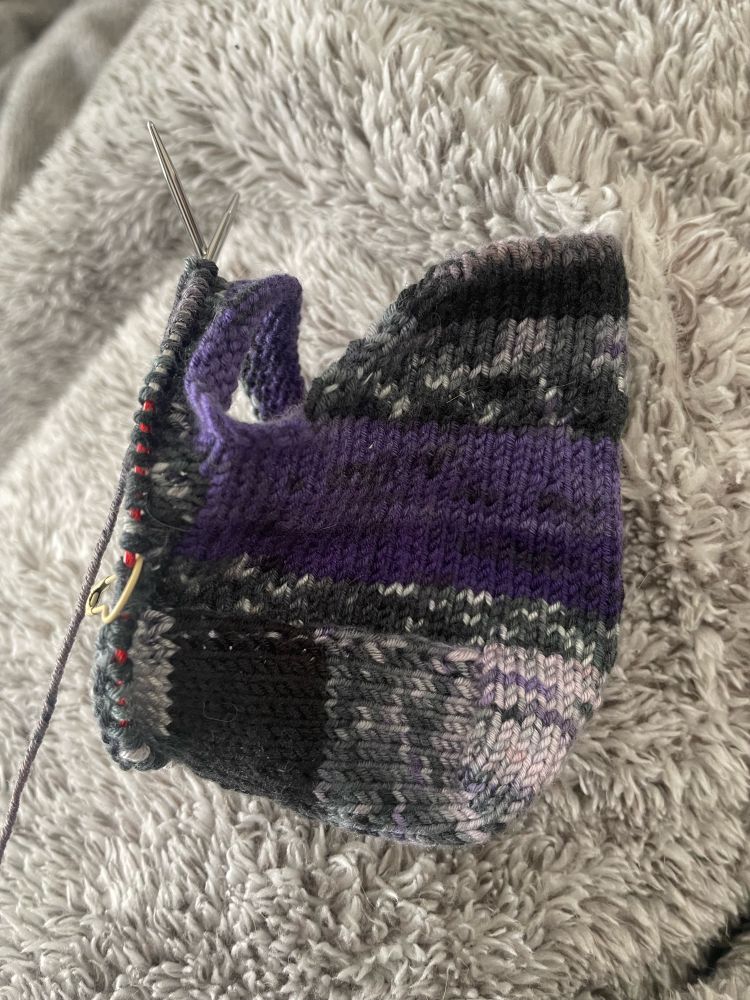 The beginning of a purple handknit sock on the needles, resting on a furry blanket. 