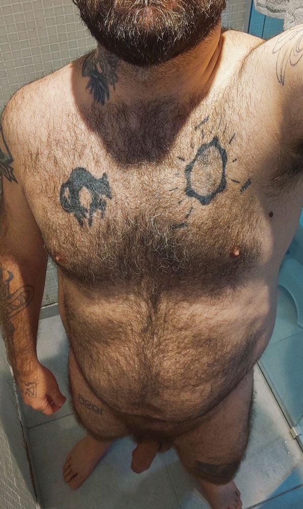 Eu nu no box do banheiro, de pé, tirando foto visto de cima. Sou um homem cis branco de 38 anos, com o corpo um pouco acima do peso ideal, peludo e tatuado. Minha barba grisalha pode ser vista parcialmente na parte superior da foto que mostra até meus pés.
