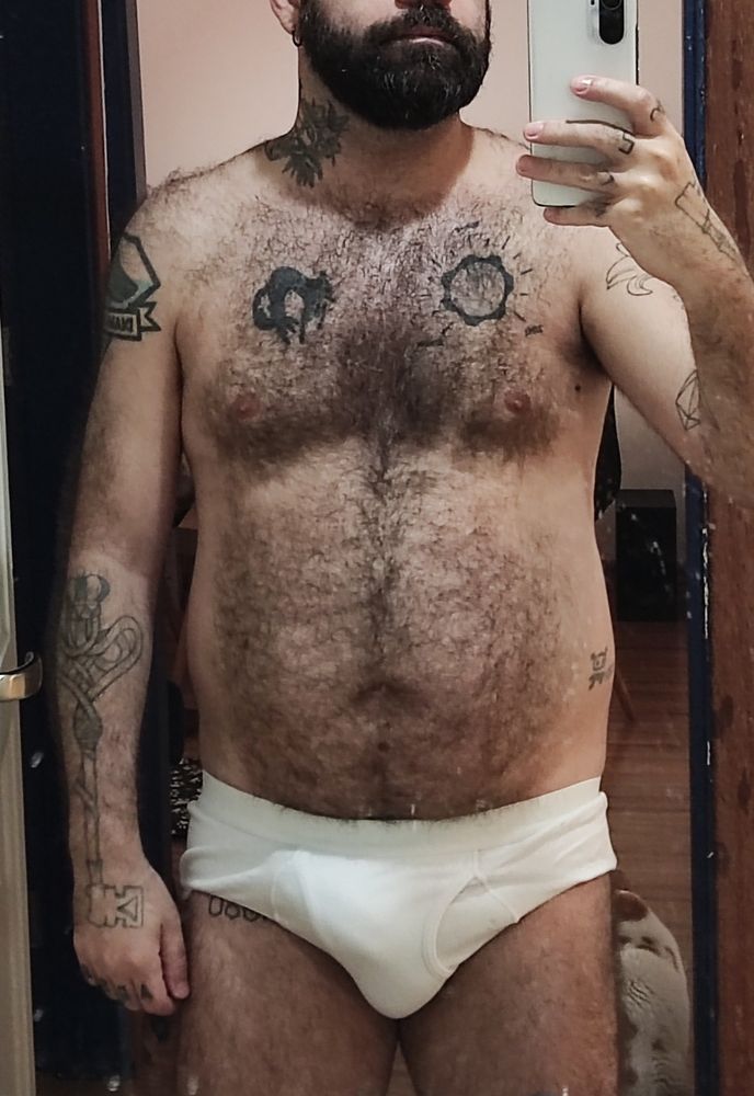 Me standing in front of a mirror, wearing just white briefs, with an erection bulge showing. My body is hairy, slightly beefy and has various tattoos on my chest, arms, hands, neck and legs.