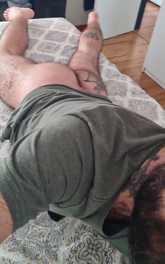 38 y.o. white male laying stomach down on a bed, wearing just a t-shirt, taking a selfie. His face is partially hidden and his naked butt and legs are visible. His beard is black with some white hair, his butt and legs are furry and tattooed.
