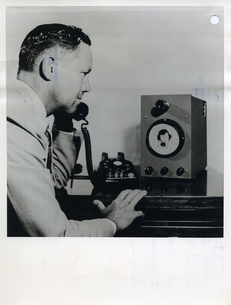 black and white photograph of a man holding a telephone receiver to his ear while looking at a box with a miniature screen showing a live feed of the caller on the other end. 