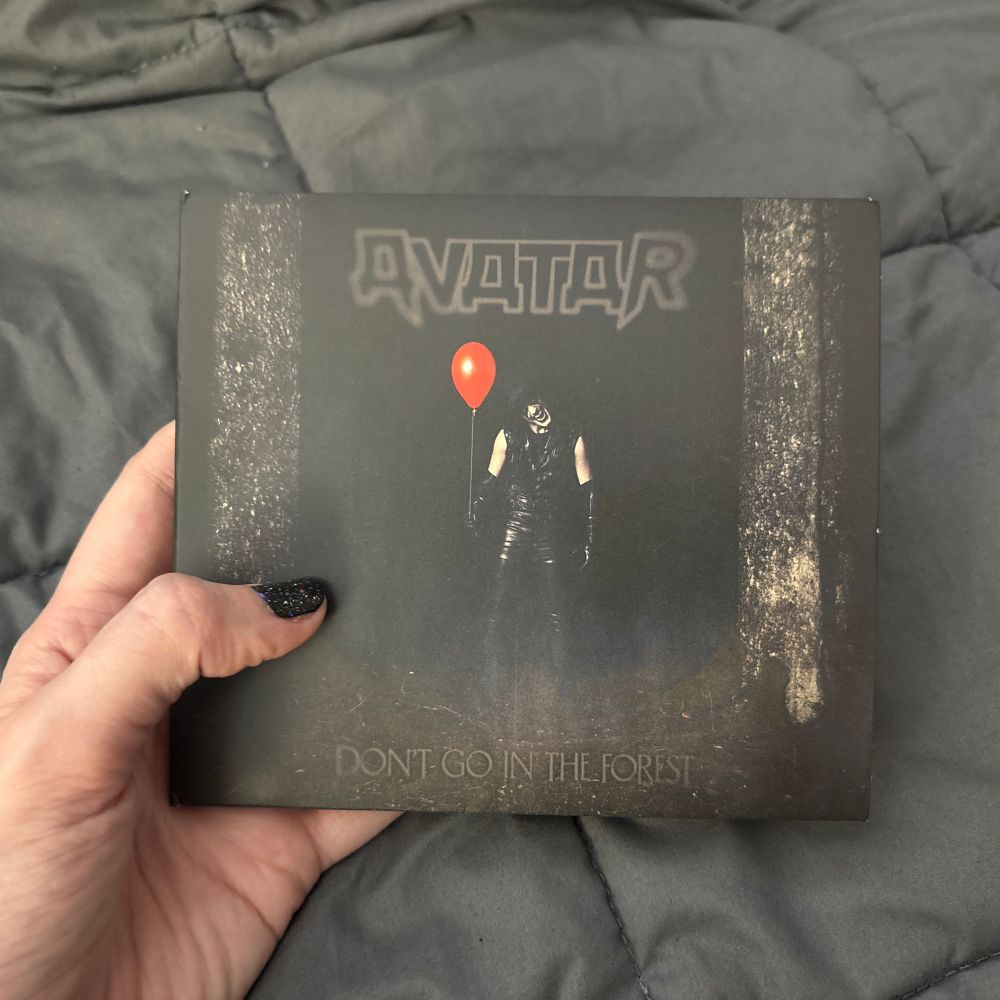 A photo of a hand holding up a CD copy of Avatar’s latest album, “Don’t Go In The Forest” - the cover depicts the lead singer Johannes standing in front of a black background dressed in black pleather and his signature clown like face paint. He is making a creepy smiling face and holds a single red balloon.